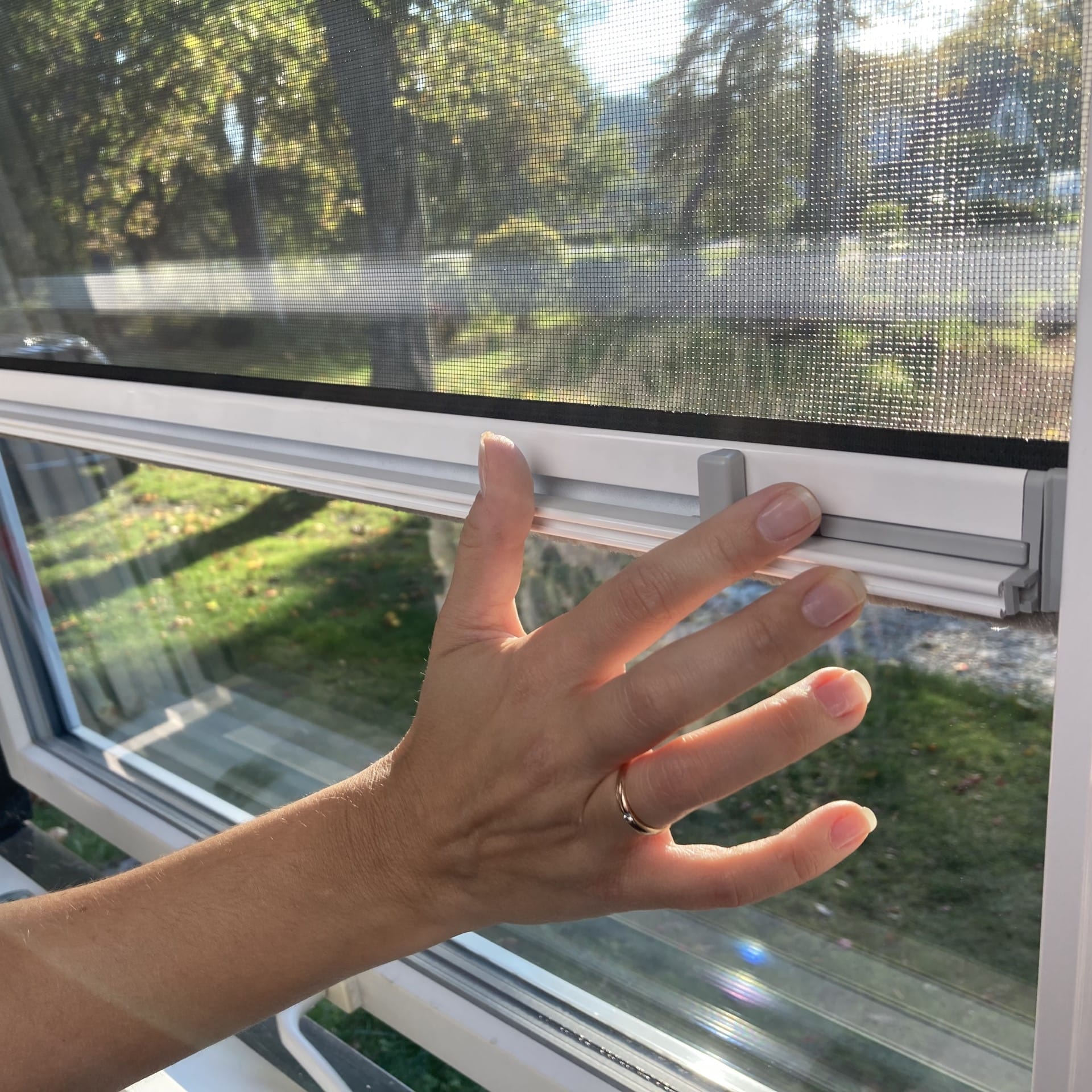 A hand with a ring opens a window by sliding the latch upward on a sunny day, with trees and grass visible outside.
