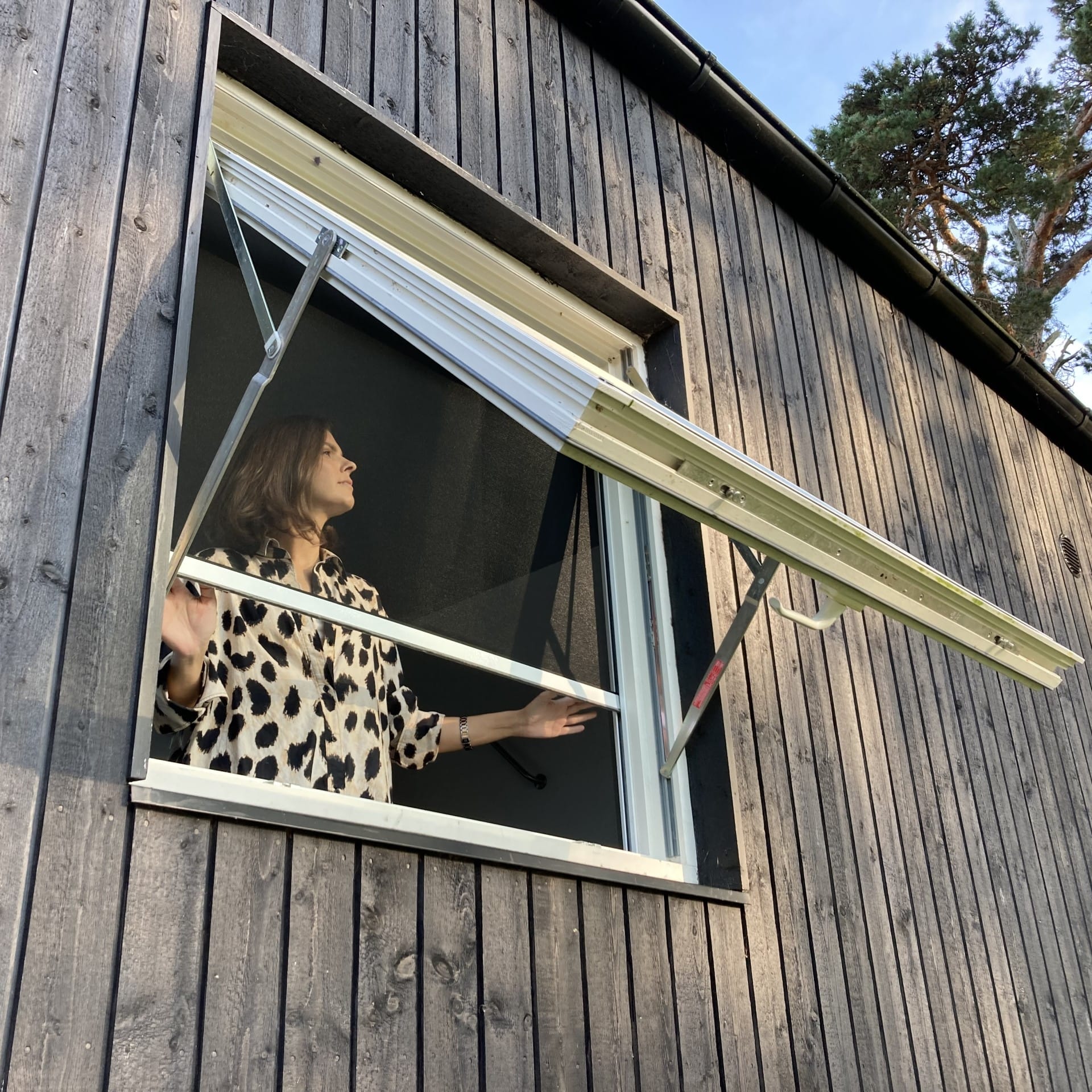 Sieviete kreklā ar leoparda raksta apdruku stāv pie atvērta koka ēkas loga un lūkojas ārā.