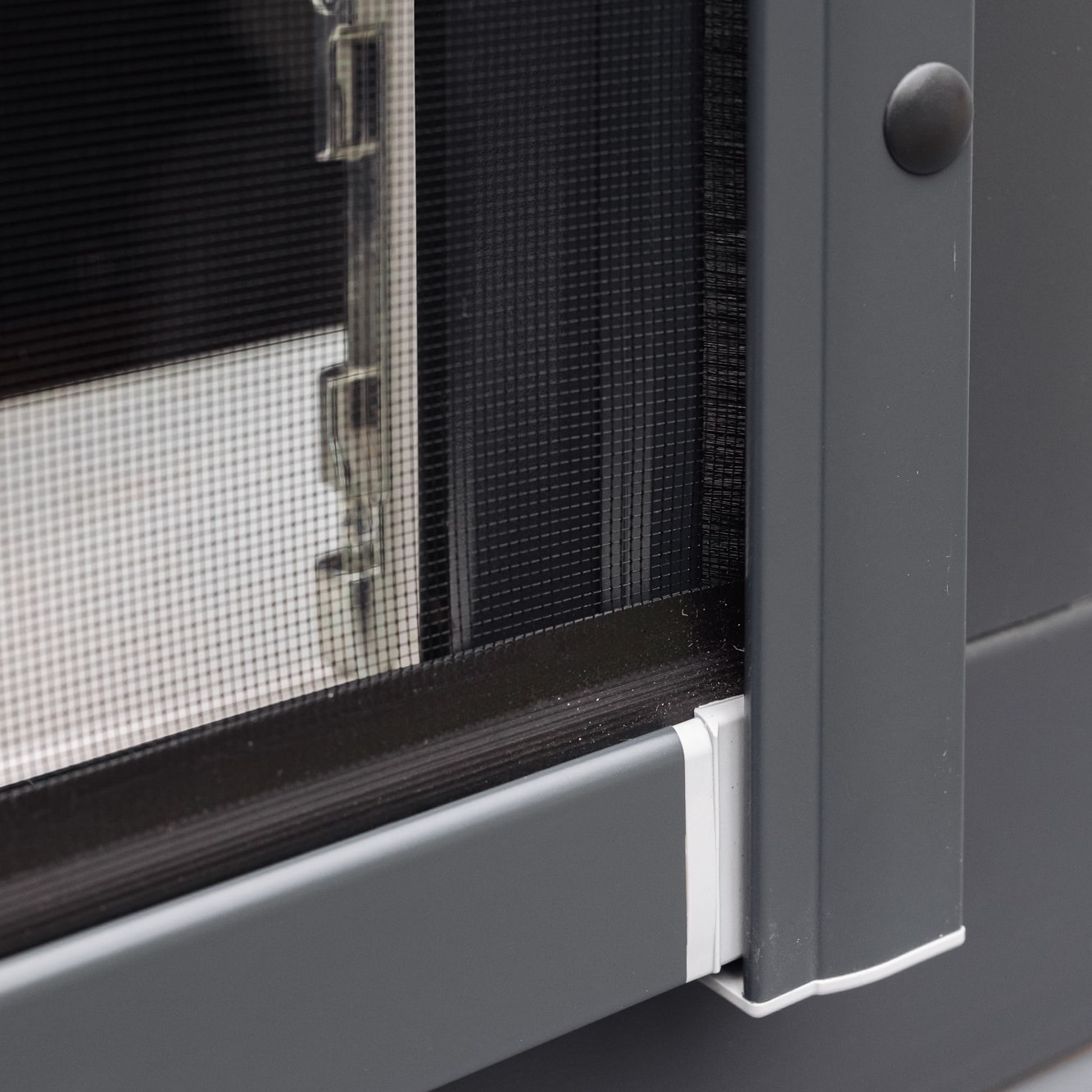 Lähikuva Hyttysverkko ikkunaan - rullaverkko ikkunaruudun nurkasta, jossa on harmaa kehys ja valkoinen muovinen liitospala.