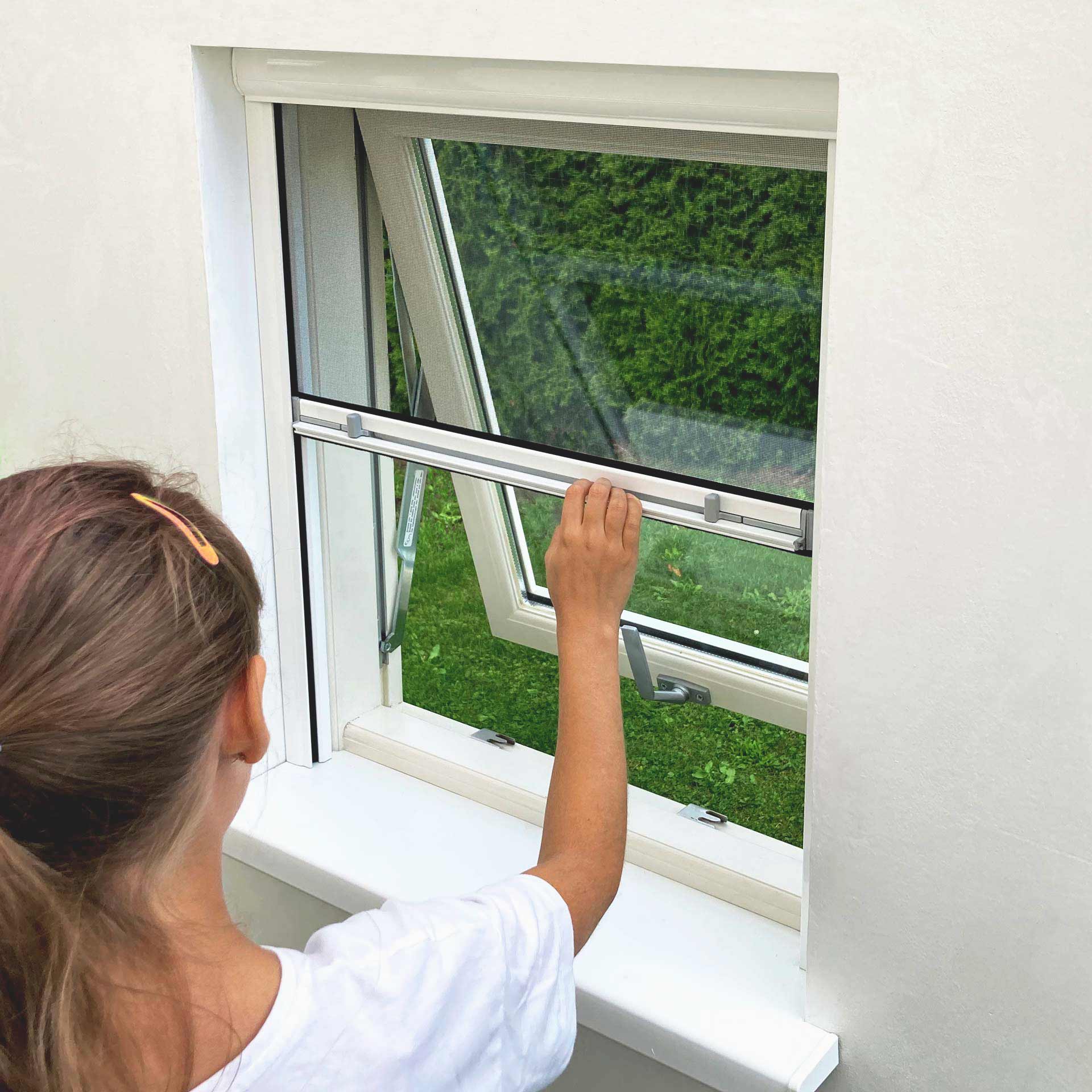 Valkoiseen paitaan pukeutunut henkilö avaa kaksilasisen ikkunan Hyttysverkko ikkunaan - rullaverkko; vihreä pensasaita näkyy ulkona.