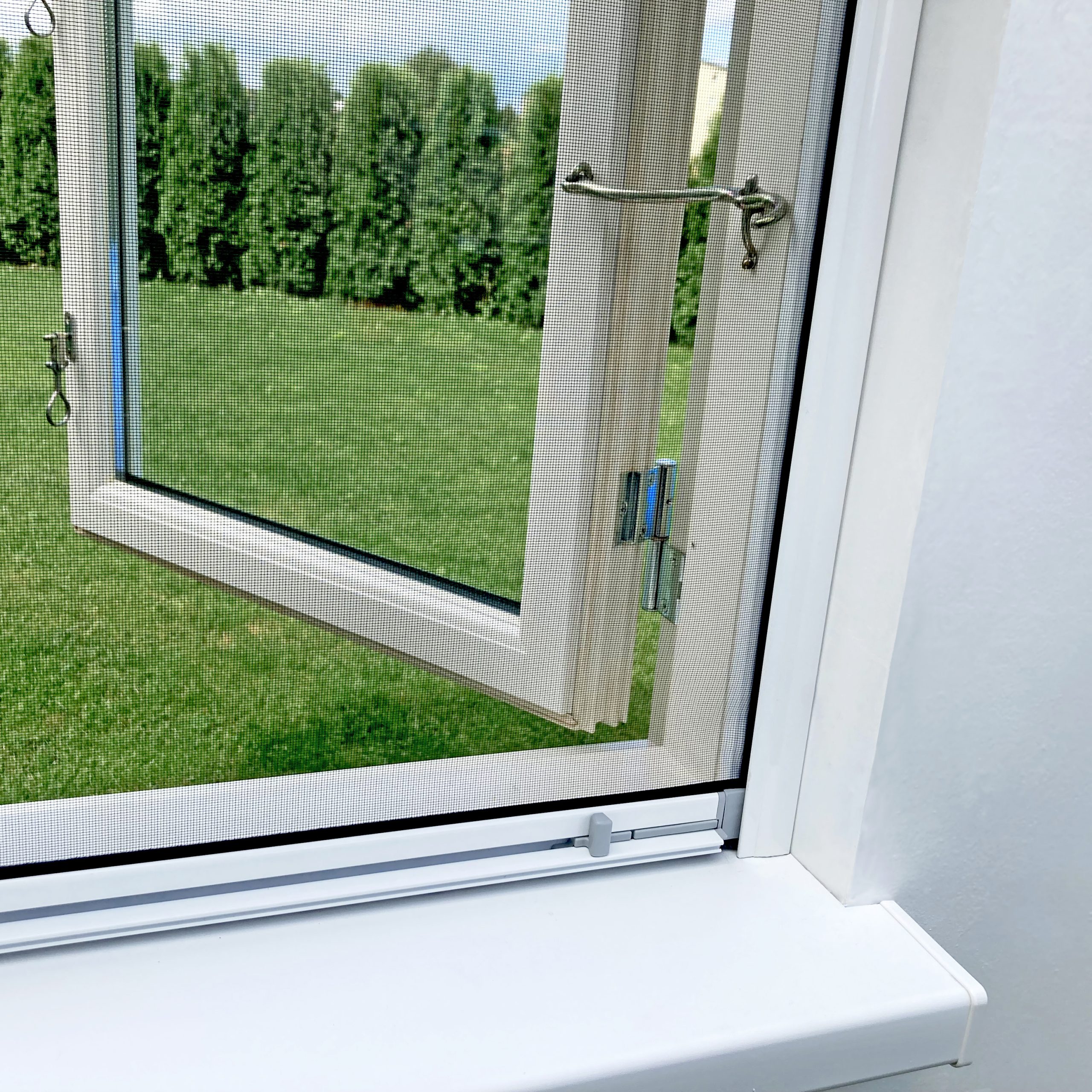 Avoin valkopuitteinen ikkuna Hyttysverkko ikkunaan - rullaverkko, josta näkymät vihreälle nurmikolle ja korkeille puille.