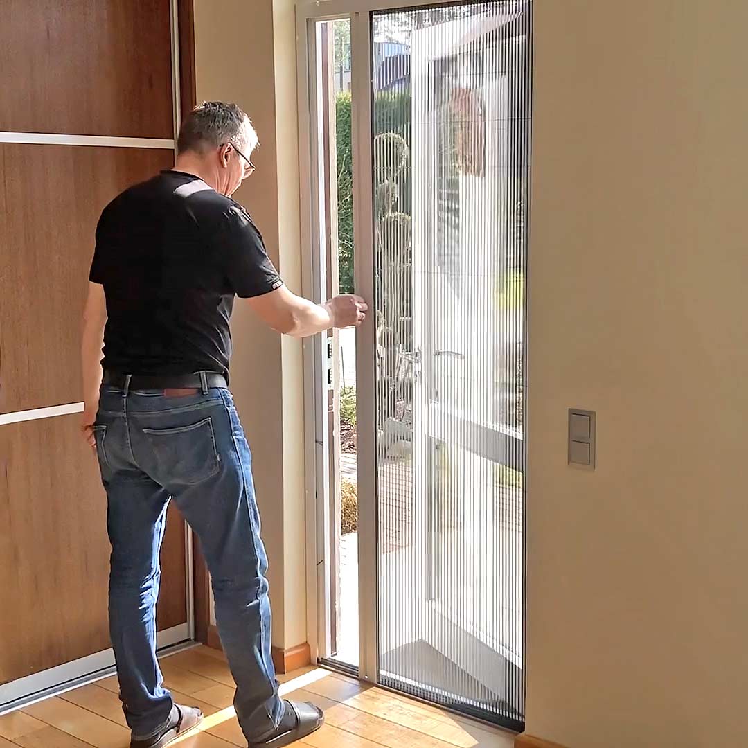 Ein Mann in Freizeitkleidung öffnet ein Fliegengitter Tür - faltbar, um aus einem sonnenbeschienenen Raum nach draußen zu treten.
