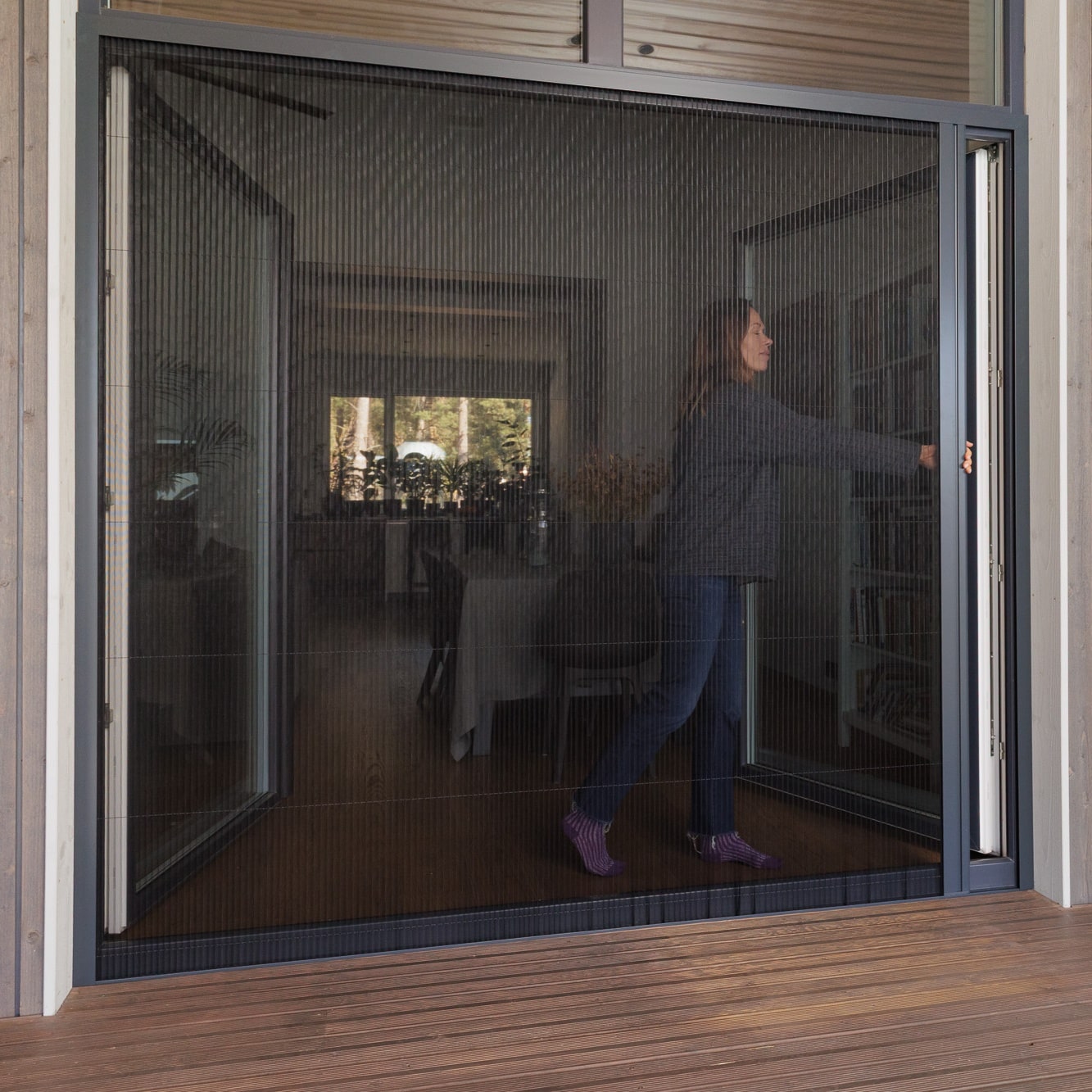 A woman opens a foldable insect screen door from a wooden deck into a modern, well-lit living space.