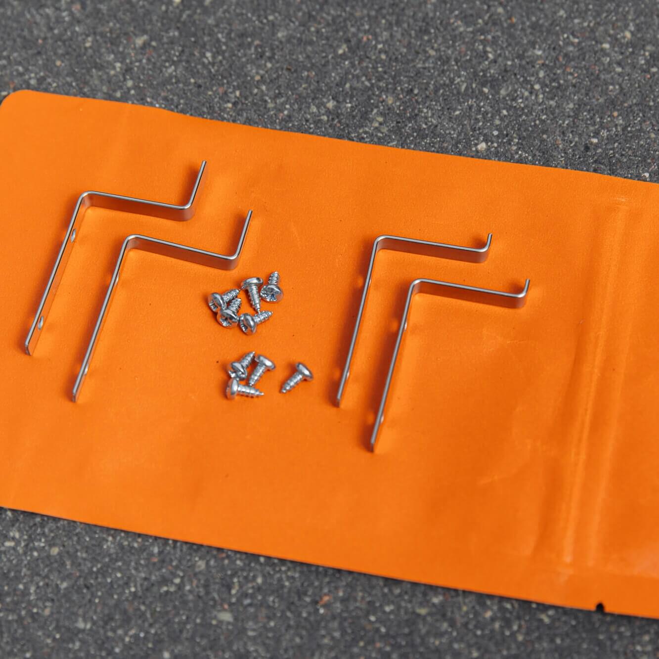 Three metal L-brackets and screws for the Insect screen window – in frame are displayed on an orange surface with a gray backdrop.