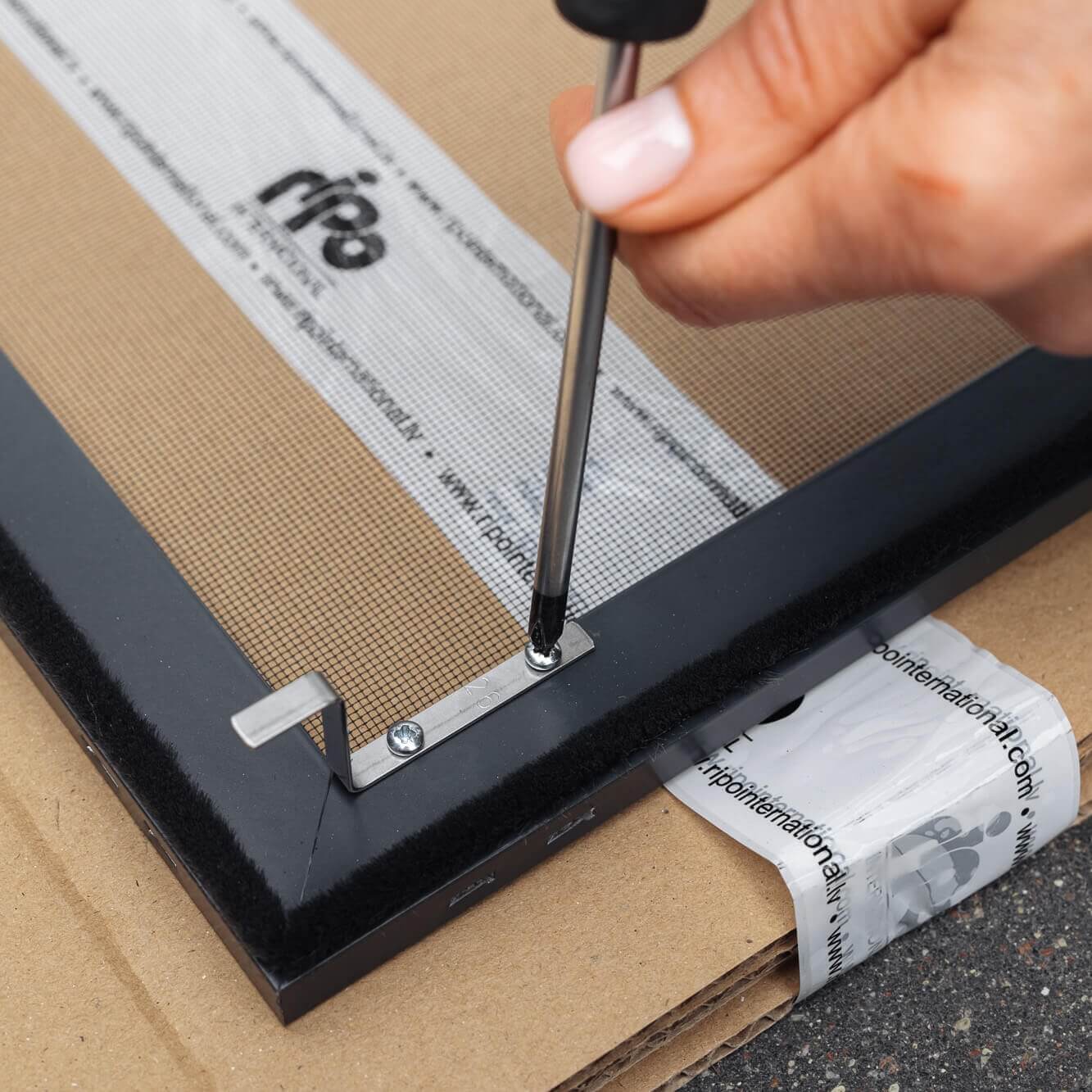 Henkilö kiinnittää ruuvimeisselillä metallikannattimen Hyttysverkko ikkunaan - kehysmalli kartongin kulmaan.